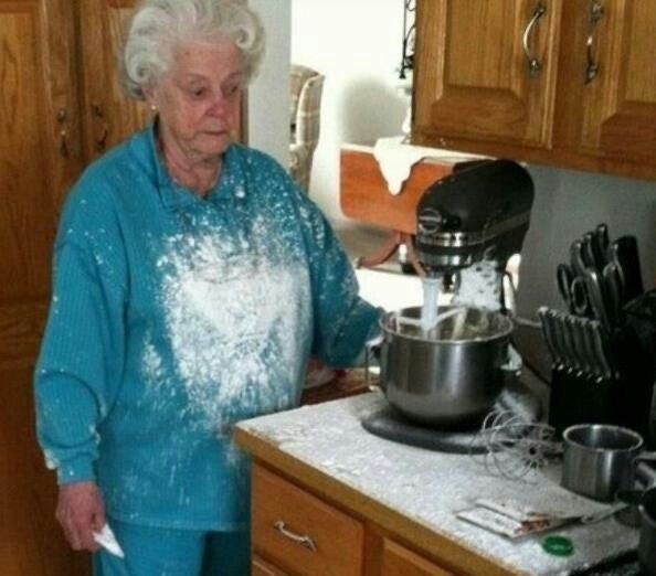 a grandmother in a teal sweatsuit standing at her stand mixer, she and the whole kitchen covered in flour