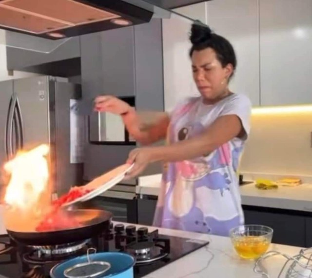 a woman tipping a plate of chopped tomatoes into a frying pan that has burst into flames, leaning away and wincing