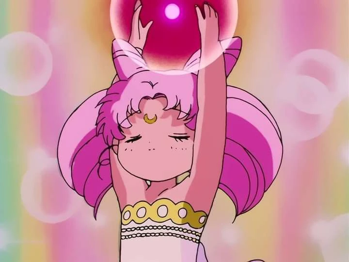 Chibiusa in a pale gown with a gold band, eyes closed and both arms raised, holding the Pink Moon Crystal above her head as it glows against a haze of pastel light