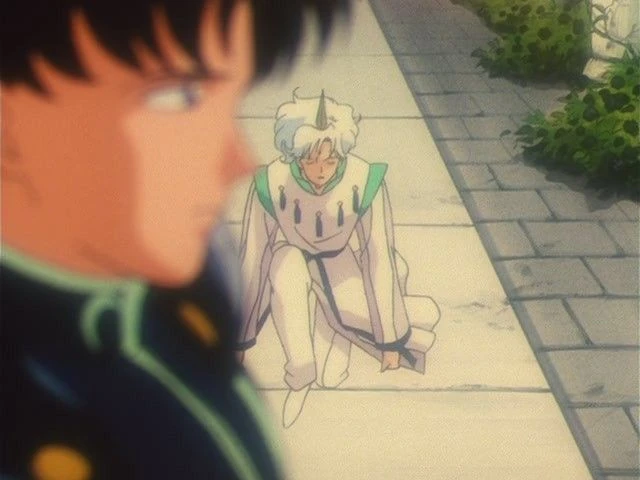Mamoru out of focus in the foreground, Helios in white robes kneeling on the stone path behind him