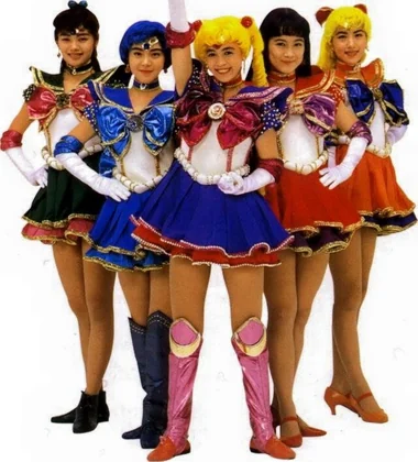 Five musical performers in beaded stage costumes, hands on hips, Sailor Moon centre with one arm raised
