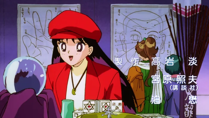 Rei in a red beret and jacket behind a fortune-telling table, crystal ball at her elbow, palmistry and face-reading charts pinned up behind her