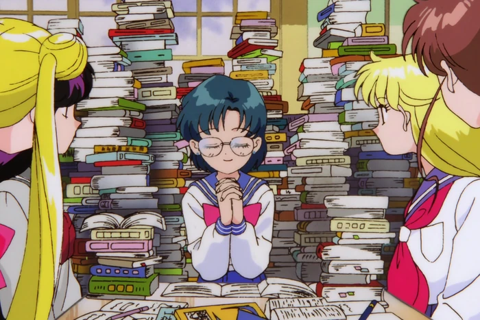 Ami in her reading glasses at the centre of towering stacks of books, hands clasped and eyes lowered, with Usagi, Rei, Minako and Makoto gathered round the table looking at her