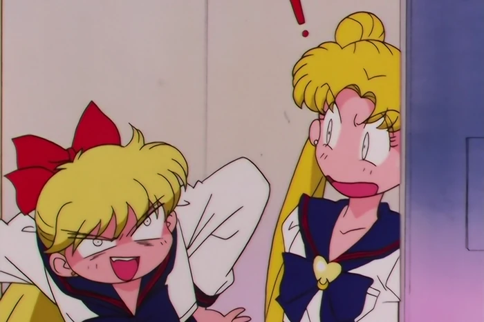 Minako grinning with her tongue out and an elbow flung out to the side, while Usagi presses herself back against the wall with an exclamation mark over her head