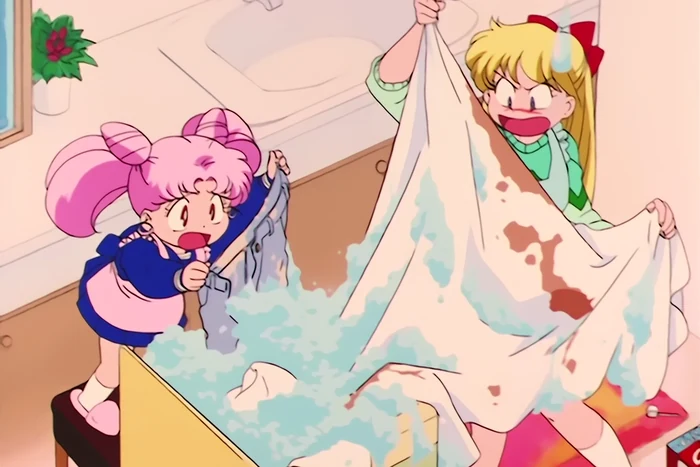 A flooded bathroom: Minako in an apron holding up a bedsheet blotched with brown stains, furious, while Chibi-Usa stands on a stool wringing out a grey garment over a tub boiling with suds