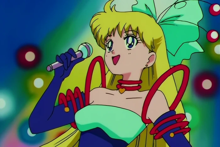 Minako on stage in a strapless mint and navy gown with red cord trim, a microphone in one hand, singing under coloured lights