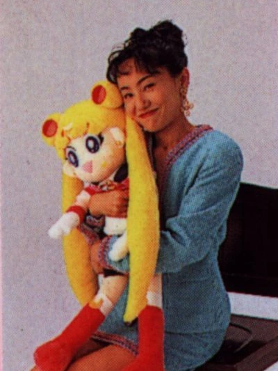 Naoko Takeuchi in a pale blue suit, seated on a black chair, hugging a large Sailor Moon plush doll cheek to cheek