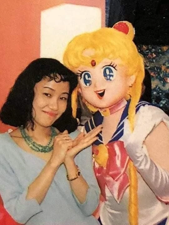 Naoko Takeuchi in a pale blue top and green beaded necklace, smiling beside a costumed Sailor Moon character, with Sailor Moon dolls on a shelf behind