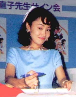 Naoko Takeuchi in a blue dress at a signing table, pen in hand, Sailor Moon posters behind her