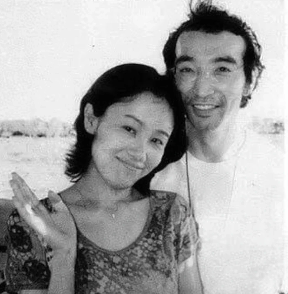 Black and white photograph of Naoko Takeuchi and Yoshihiro Togashi smiling together outdoors by the water, her hand raised in a wave